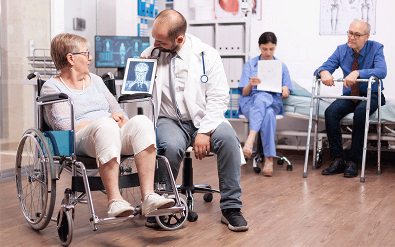 Older woman sitting in wheelchair in a physical therapy setting is accompanied by a doctor who is showing her medical imaging